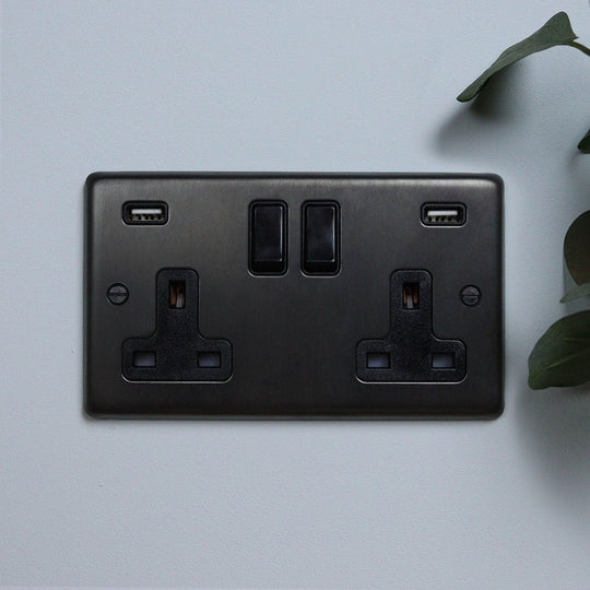 A dark bronze double socket with USB  against a light blue background with a green plant to the side.