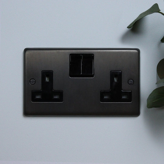 A dark bronze double socket outlet against a light blue background with a green plant to the side.